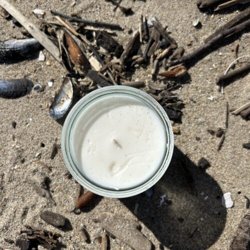 An overhead view of an unlit, white candle in a glass jar. The candle is on the sand and bits of ocean debris like shells, sticks, and seaweed are scattered around the sand.