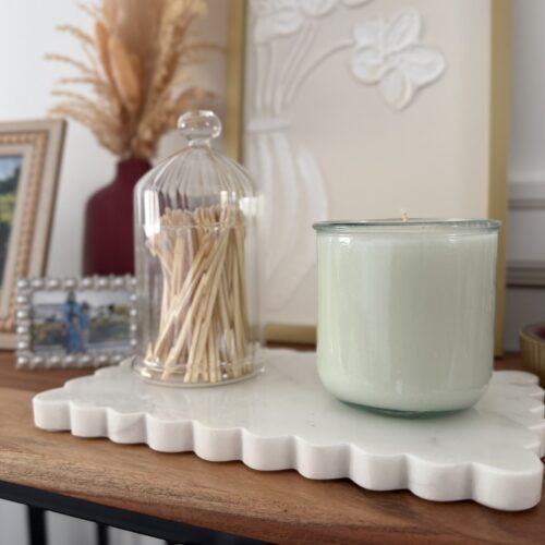 A Relume candle sits on a marble platter atop a wood table. Framed pictures of Relume staff, a cloche of candles, and a vase with decorative tan grasses are in the background.