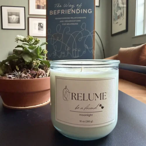A limited edition Relume "be a friend" candle sits on a coffee table next to a plant. The Way of Befriending book, is also on the table in the background.